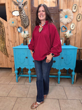 Load image into Gallery viewer, A woman modeling a dark and light red vertically striped long-sleeve blouse with puff sleeves, paired with dark blue jeans and brown sandals, standing in front of a rustic wood-paneled wall with a turquoise sideboard.