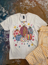 Load image into Gallery viewer, A white short-sleeve graphic T-shirt laid flat on a textured brown and blue surface, featuring a colorful autumn pumpkin print with leopard, flowers, stars and swirl patterns. A silver chain necklace is draped over the shoulders, and yellow-and-white gingham pants are partially visible at the lower right.
