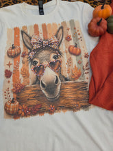 Load image into Gallery viewer, A white crewneck t-shirt featuring a colorful graphic print of a donkey wearing heart-shaped sunglasses and a leopard print bow, surrounded by autumn pumpkins, leaves, and stars. The shirt is styled flat-lay with small pumpkins and a rust-colored fabric on a leopard-print surface.
