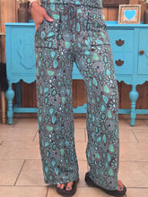 Load image into Gallery viewer, A person wearing loose, wide-leg drawstring pants with an intricate teal, gray, and black paisley-style pattern, standing in front of a bright turquoise cabinet.
