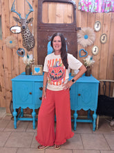 Load image into Gallery viewer, A woman stands in a decorated room wearing a white graphic t-shirt with a jack-o'-lantern print and bright dark orange wide-leg ruffled pants. She is posed in front of a blue cabinet and a wooden wall decorated with various rustic ornaments.

