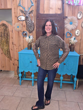 Load image into Gallery viewer, A woman modeling a leopard-print long-sleeve shirt and dark blue jeans, standing in front of a rustic wooden wall with a blue cabinet and decorative items.
