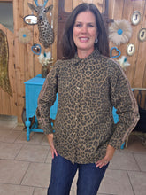 Load image into Gallery viewer, A woman wearing a brown and black leopard print button-up shirt with beige side stripes, paired with dark blue jeans, standing in a decorated rustic room with wood paneling and a blue side table.
